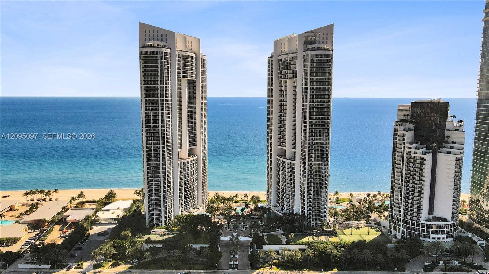 18201 Collins Avenue, #1407, Sunny Isles Beach, FL 33160 High-rise residential towers featuring expansive floor-to-ceiling glass paneling and private balconies, situated on a coastal site with direct oceanfront access and meticulously landscaped grounds