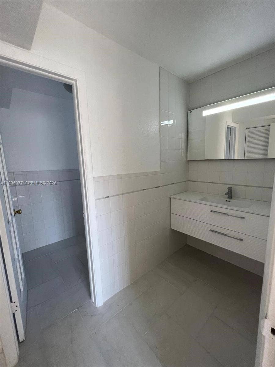 570 NW 41st Street, Miami, FL 33127 Wall-mounted vanity featuring a white composite countertop, integrated sink, and polished metal hardware, paired with a wall-spanning mirror with integrated lighting and full-height wall tiling