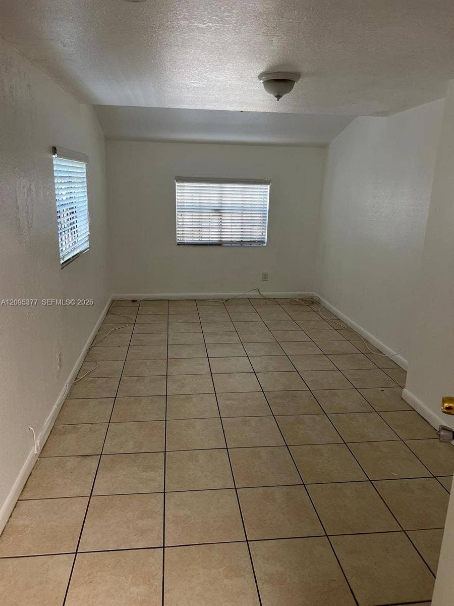 570 NW 41st Street, Miami, FL 33127 Interior room featuring large-format ceramic tile flooring, neutral wall finishes, and two windows equipped with horizontal blinds, illuminated by a flush-mount ceiling fixture
