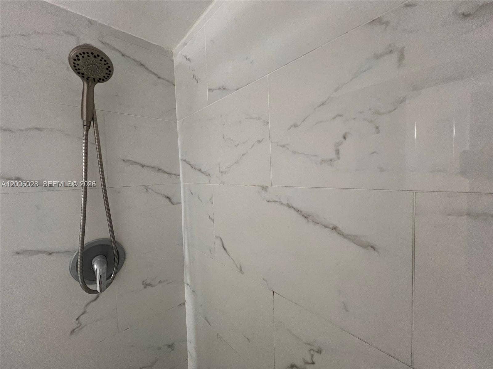 6300 SW 138th Court, #405, Miami, FL 33183 Shower surround featuring large-format, light-toned stone-finish tile with integrated veining, complemented by a metallic handheld showerhead and circular wall-mounted control valve