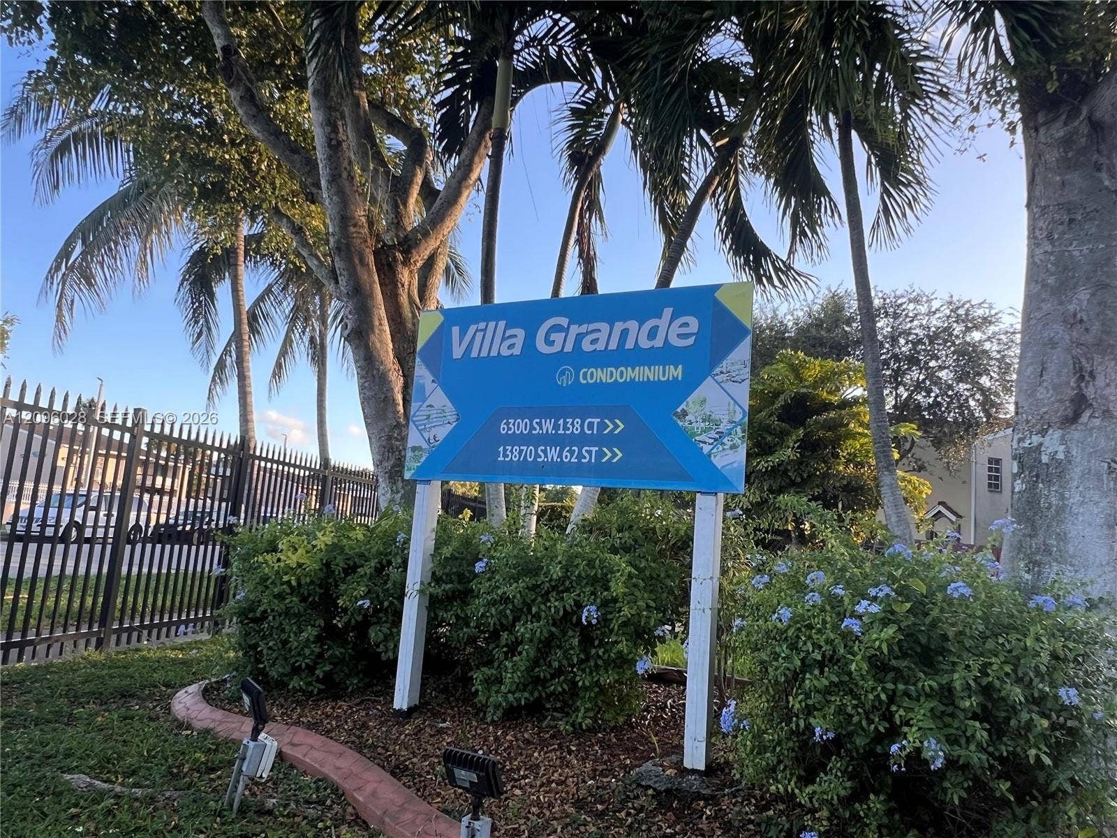 6300 SW 138th Court, #405, Miami, FL 33183 Exterior signage for Villa Grande Condominium featuring mature palm trees, perimeter metal fencing, and landscaped shrubbery