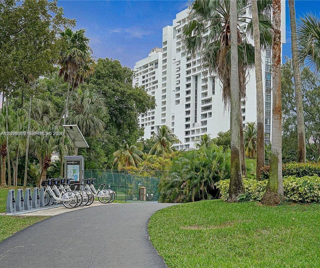 20355 NE 34th Court, #1925, Aventura, FL 33180 High-rise residential exterior featuring a multi-story white facade with recessed balconies, paved pathways, mature palm trees, landscaped greenery, and a bicycle-sharing station