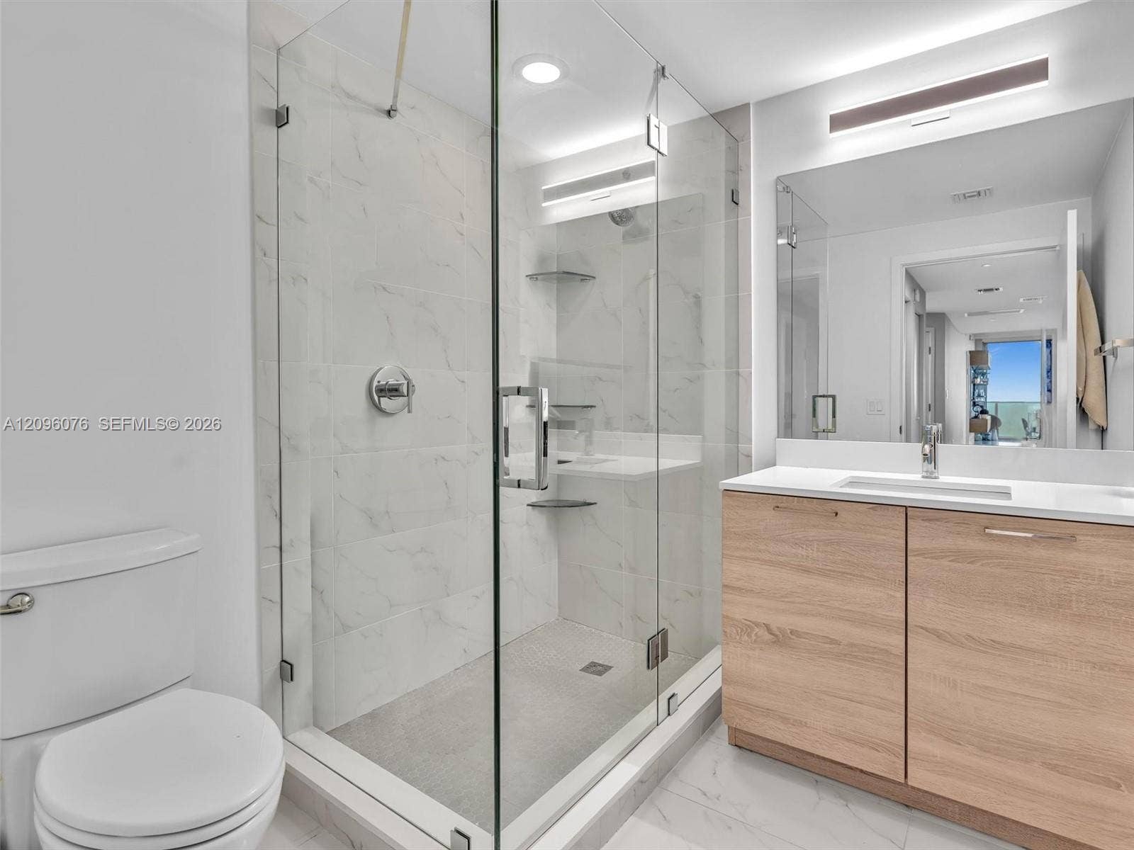 16385 Biscayne Boulevard, #1101, North Miami Beach, FL 33160 Walk-in glass-enclosed shower with stone-finish tile surround, light wood-finish cabinetry, white countertop with integrated sink, and large vanity mirror with wall-mounted lighting