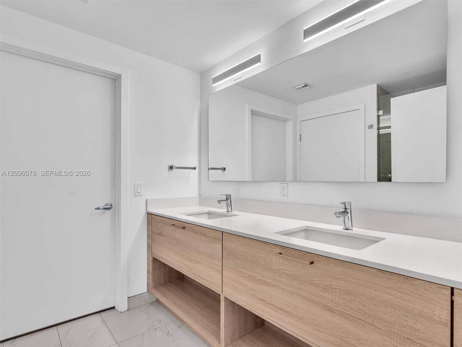 16385 Biscayne Boulevard, #1101, North Miami Beach, FL 33160 Dual-sink vanity featuring a light wood-finish cabinet with open shelving, integrated solid-surface countertop, rectangular undermount basins, and large wall-mounted mirror with recessed integrated lighting