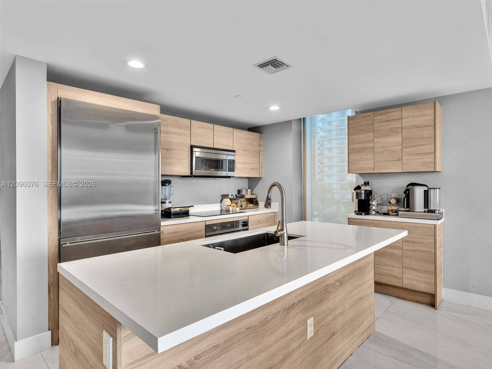16385 Biscayne Boulevard, #1101, North Miami Beach, FL 33160 Kitchen featuring wood-finish cabinetry, quartz countertops, a central island with an integrated sink, stainless steel appliances, and large-format tile flooring