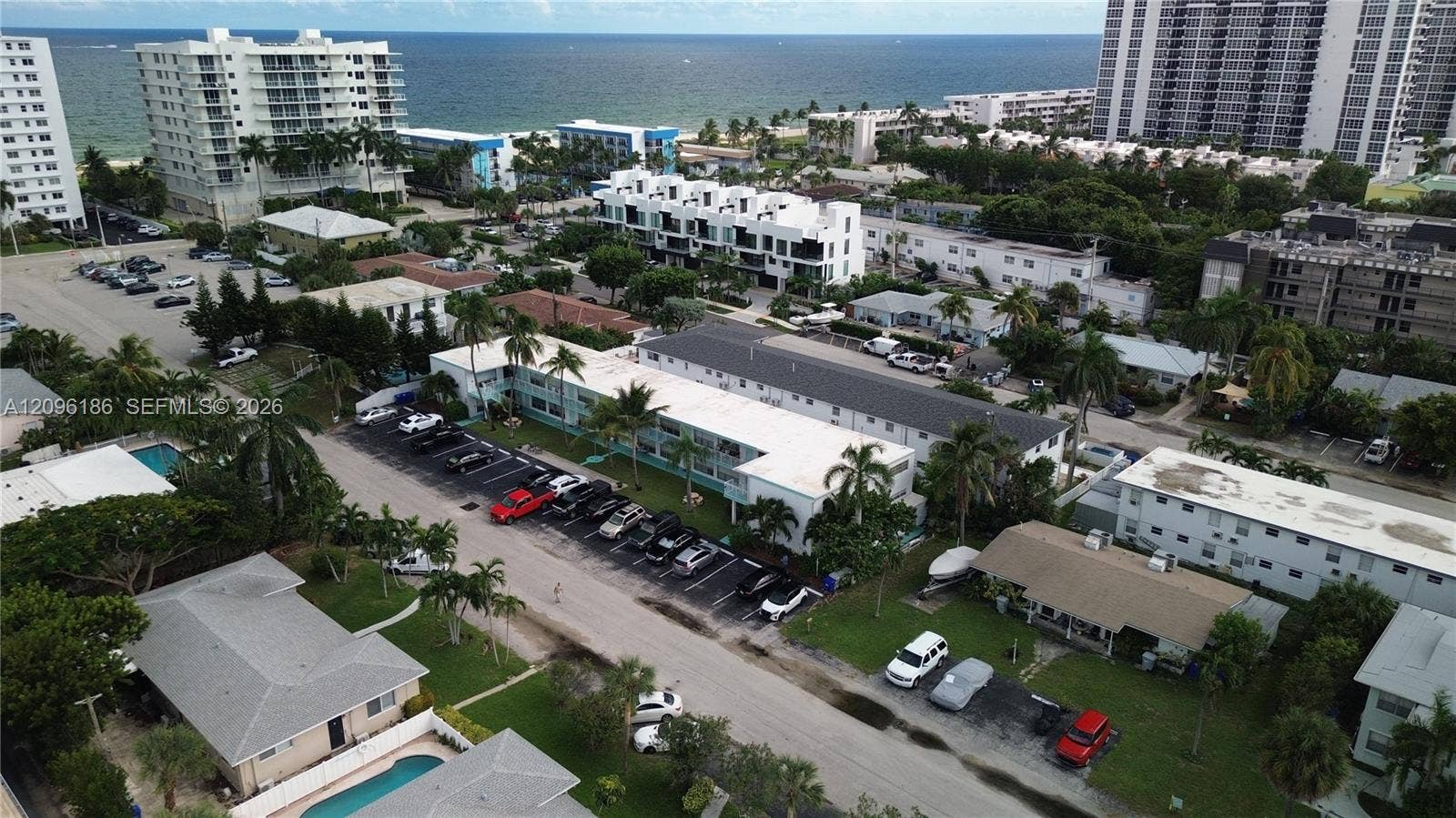 Aerial perspective of a multi-unit residential property featuring an asphalt parking lot, manicured landscaping with palm trees, and proximity to the coastline