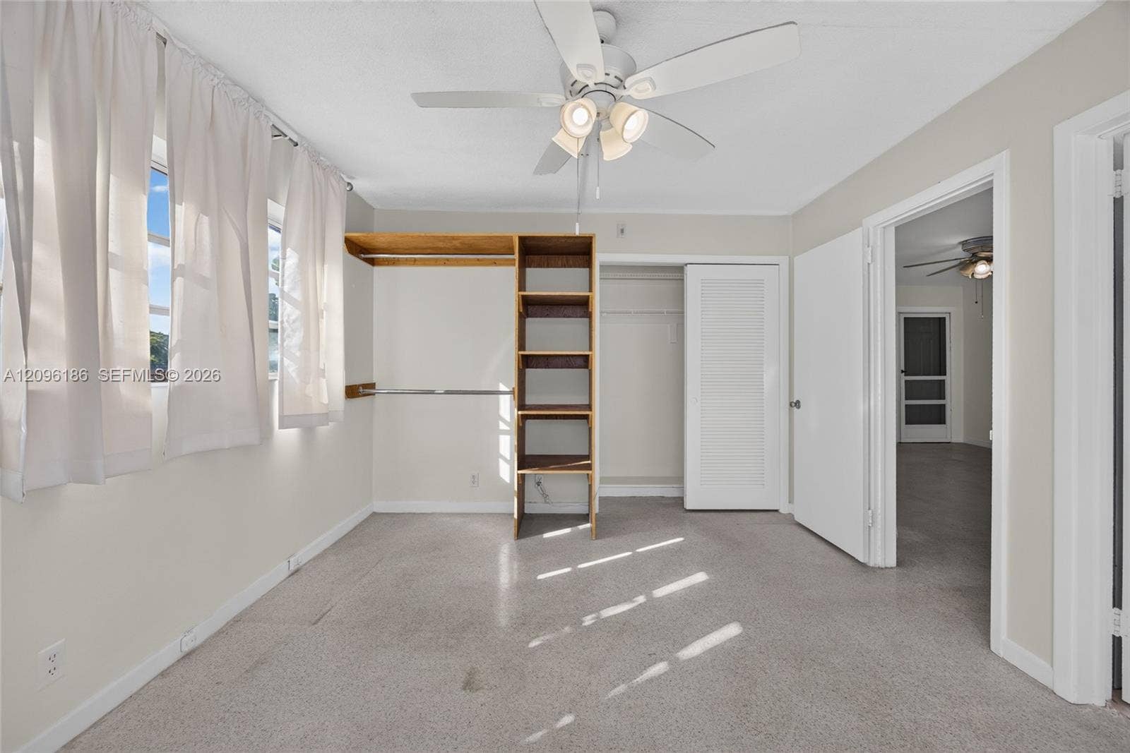 Neutral-toned room featuring a central ceiling fan with integrated lighting, open built-in shelving, carpeted flooring, and a closet with a louvered door