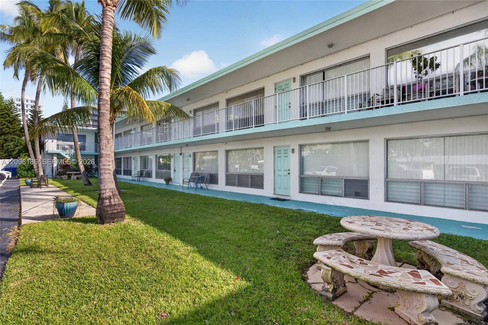 Two-story multi-unit exterior featuring white stucco siding, teal-toned trim, a second-story balcony with metal railings, and a landscaped lawn with palm trees