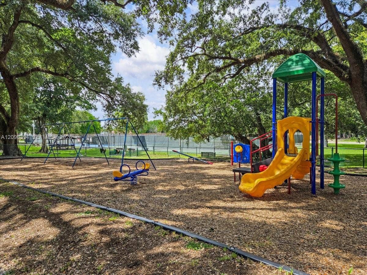 1830 Sabal Palm Drive, #105, Davie, FL 33324 Outdoor recreation area featuring a playground structure with a slide, swings, and a teeter-totter, set within a park-like space with mature trees and perimeter fencing