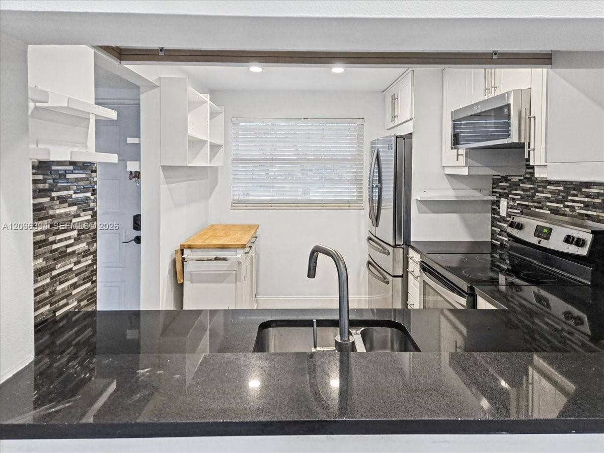 1830 Sabal Palm Drive, #105, Davie, FL 33324 Kitchen featuring black stone countertops, a stainless steel dual-basin undermount sink with a high-arch faucet, white cabinetry, a multi-tone horizontal tile backsplash, and stainless steel appliances