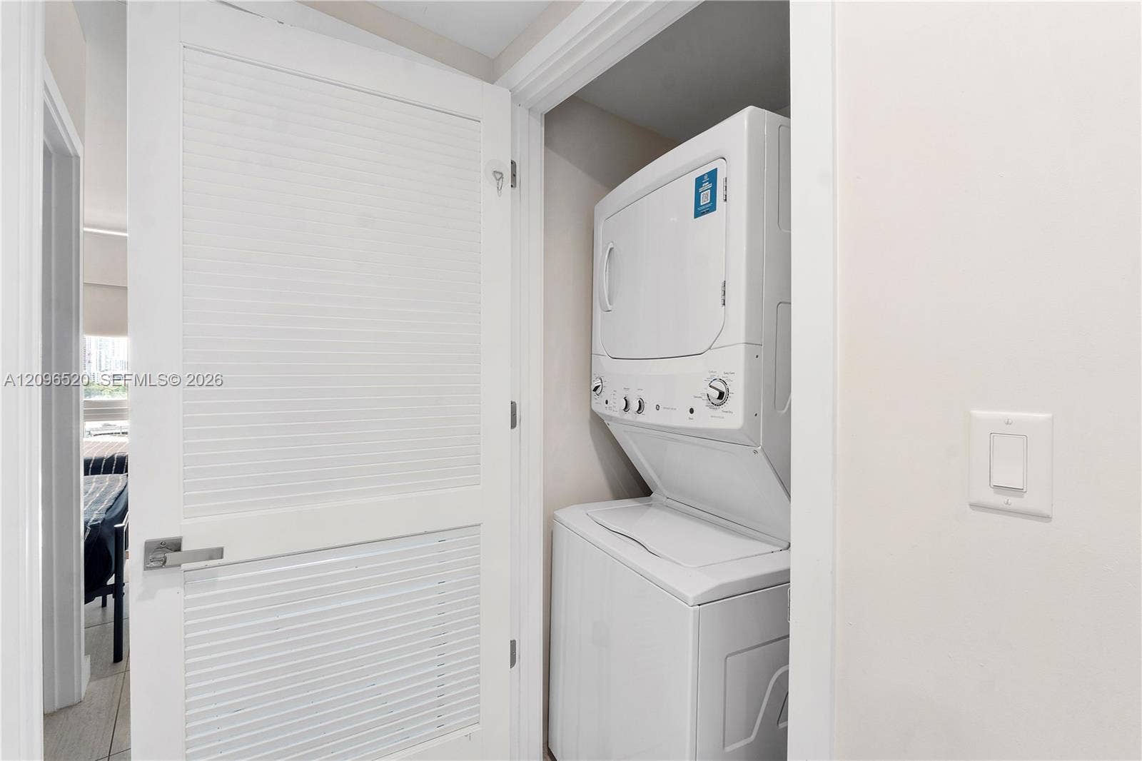 Stacked laundry unit with white finished cabinetry and a louvered door featuring a brushed metal lever handle