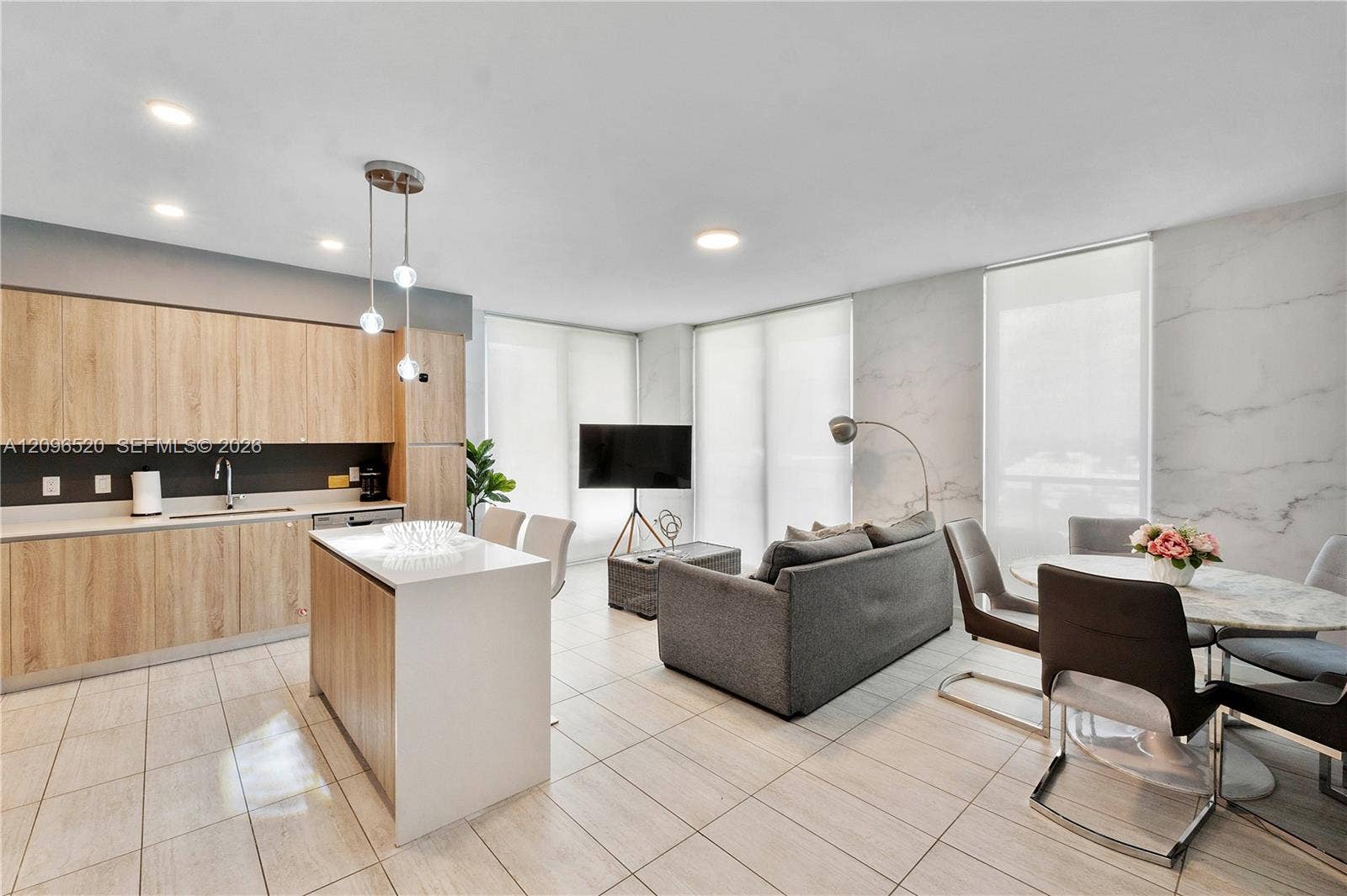 Open-concept kitchen and living area featuring wood-finish cabinetry, an island with a stone-composite countertop, light-toned tiled flooring, recessed lighting, and a large-format stone-patterned wall accent