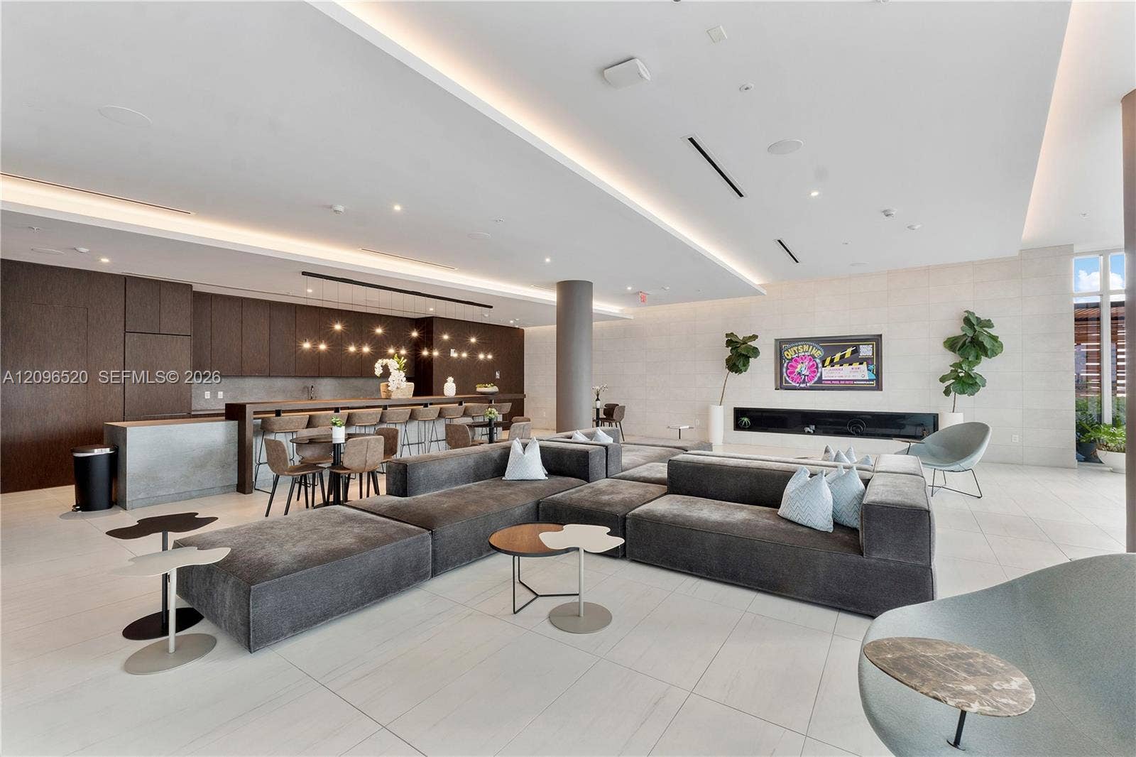 Open-concept lounge featuring large-format tile flooring, recessed cove lighting, a linear built-in fireplace, and dark-toned cabinetry with an integrated bar