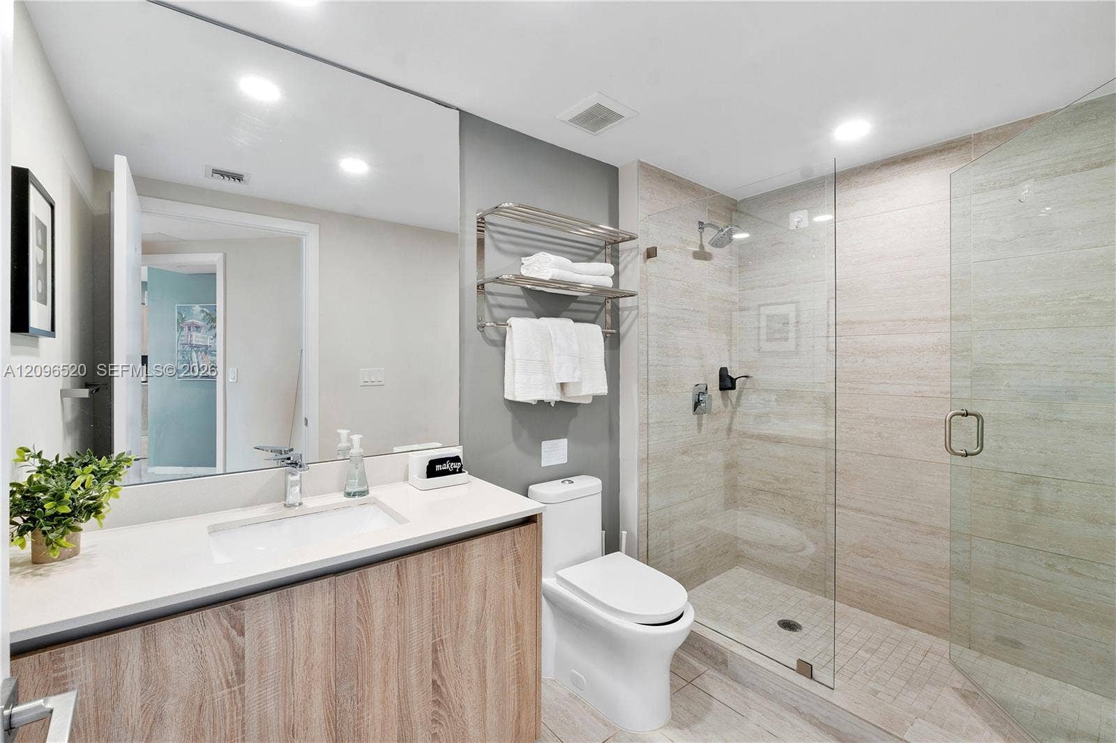 Bathroom featuring a wood-finish vanity with a quartz countertop, integrated sink, frameless glass shower enclosure with horizontal wall tile, and wall-mounted metal shelving