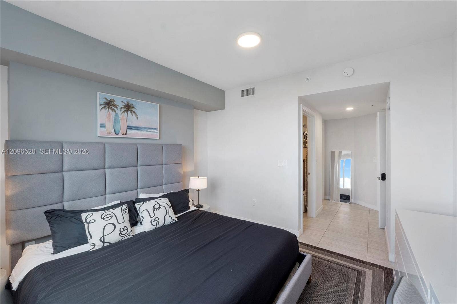 Built-in grey upholstered headboard with a geometric panel design, light-toned tile flooring, and recessed ceiling lighting