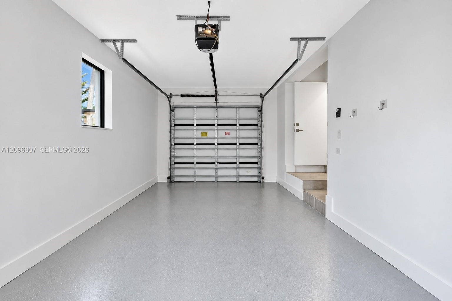 Interior garage featuring smooth white walls, a finished epoxy floor, an automatic overhead door opener, a small rectangular side window, and an interior access door with steps