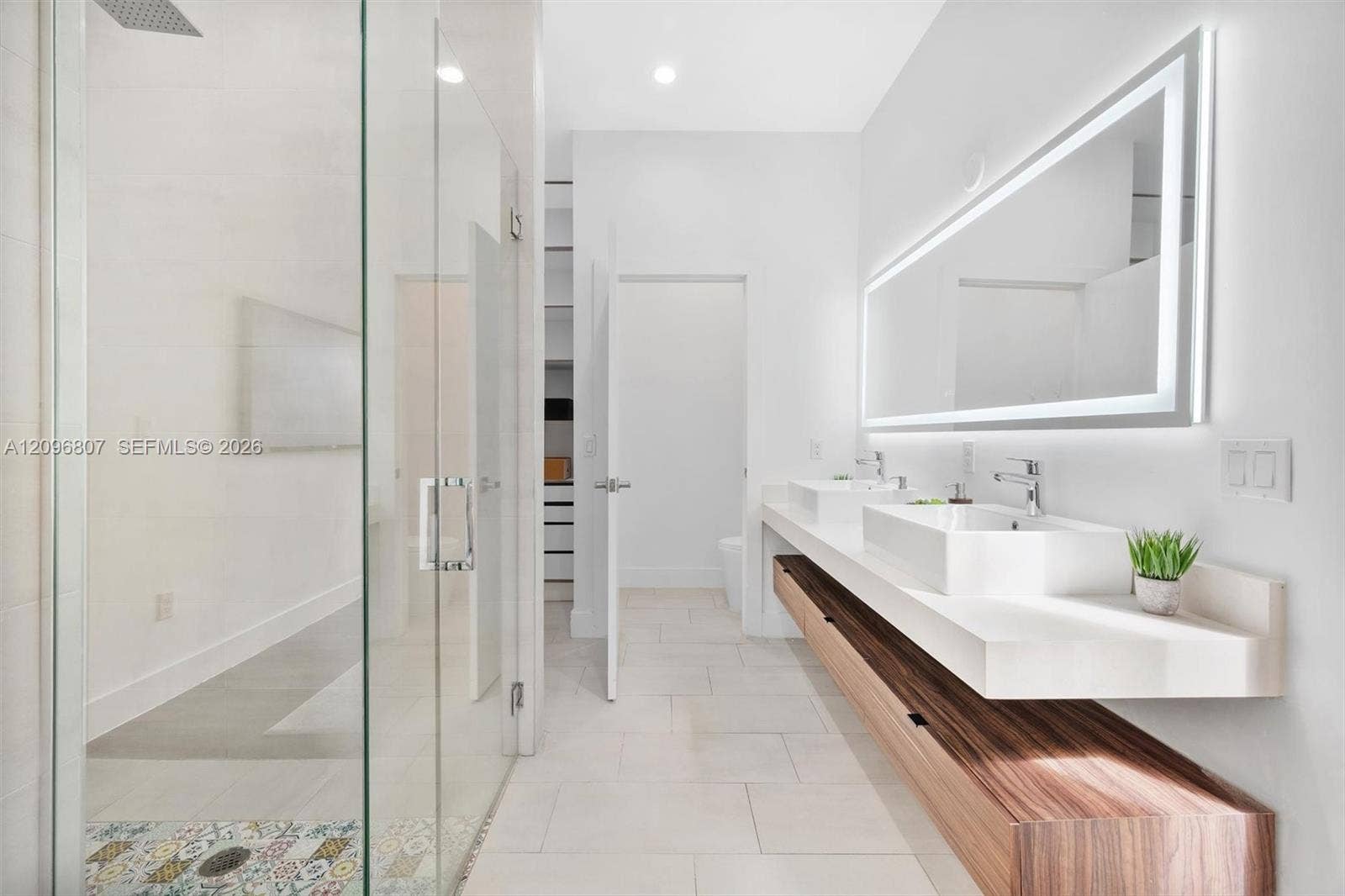 Floating dual-sink vanity with wood-finish cabinetry, quartz countertops, frameless glass walk-in shower with decorative tile accents, and an integrated LED-backlit mirror