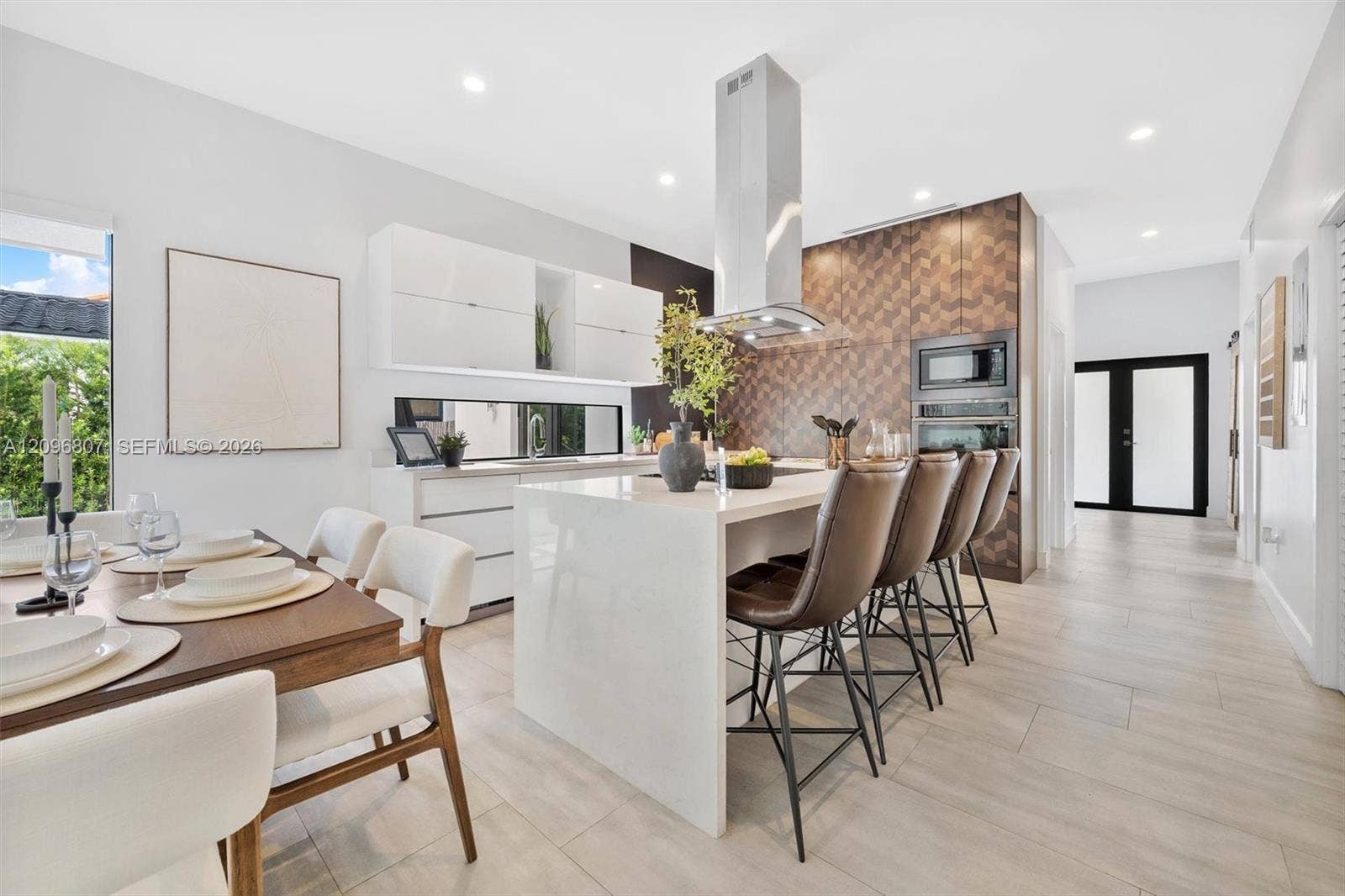 Kitchen featuring a waterfall-edge island with stone-finish countertop, high-gloss cabinetry, stainless steel range hood, patterned tile backsplash, and tile flooring