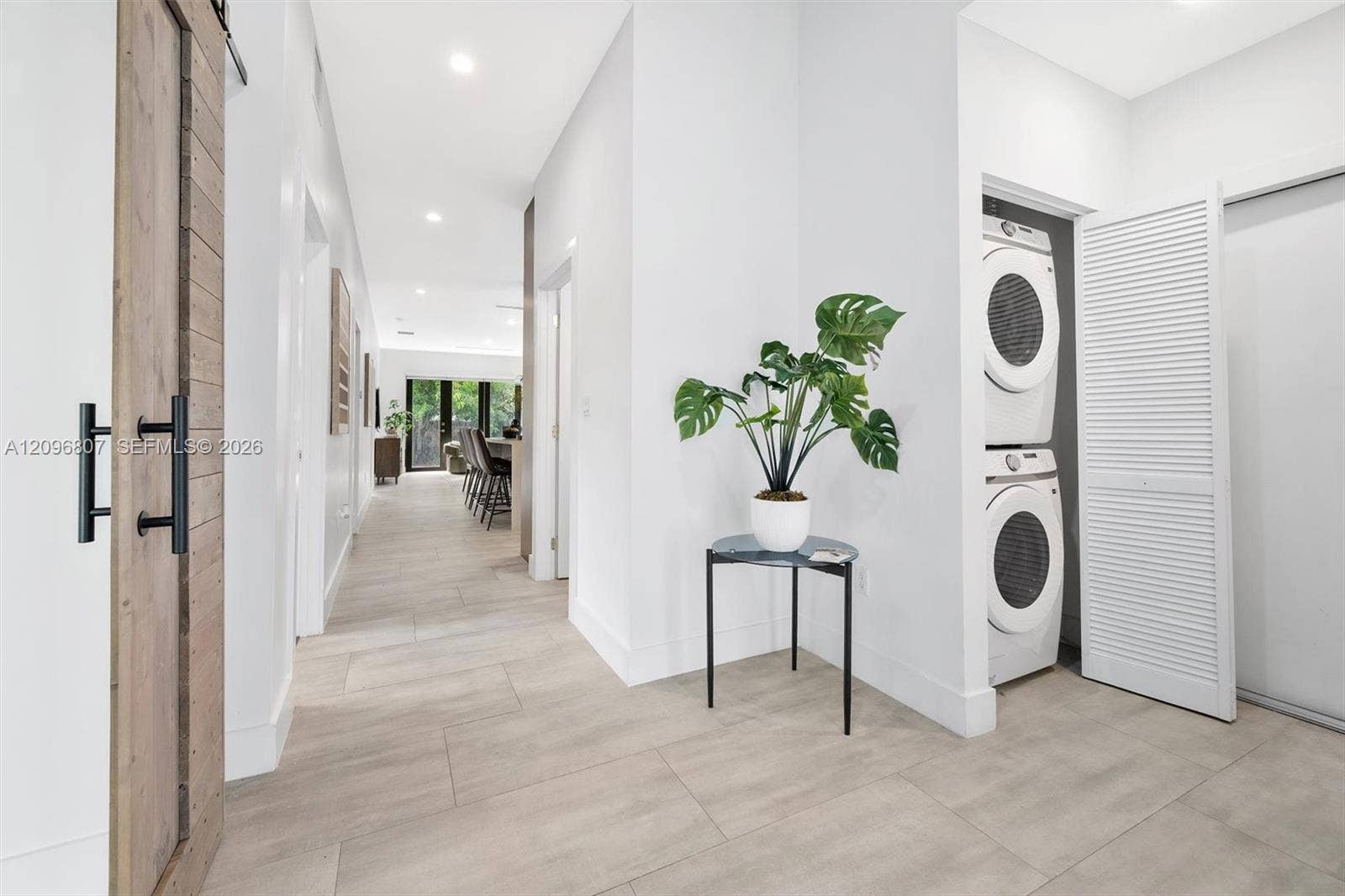 Neutral-tone tile flooring, sliding wood-finish barn door with matte black hardware, and laundry alcove featuring stacked appliances and louvered folding doors