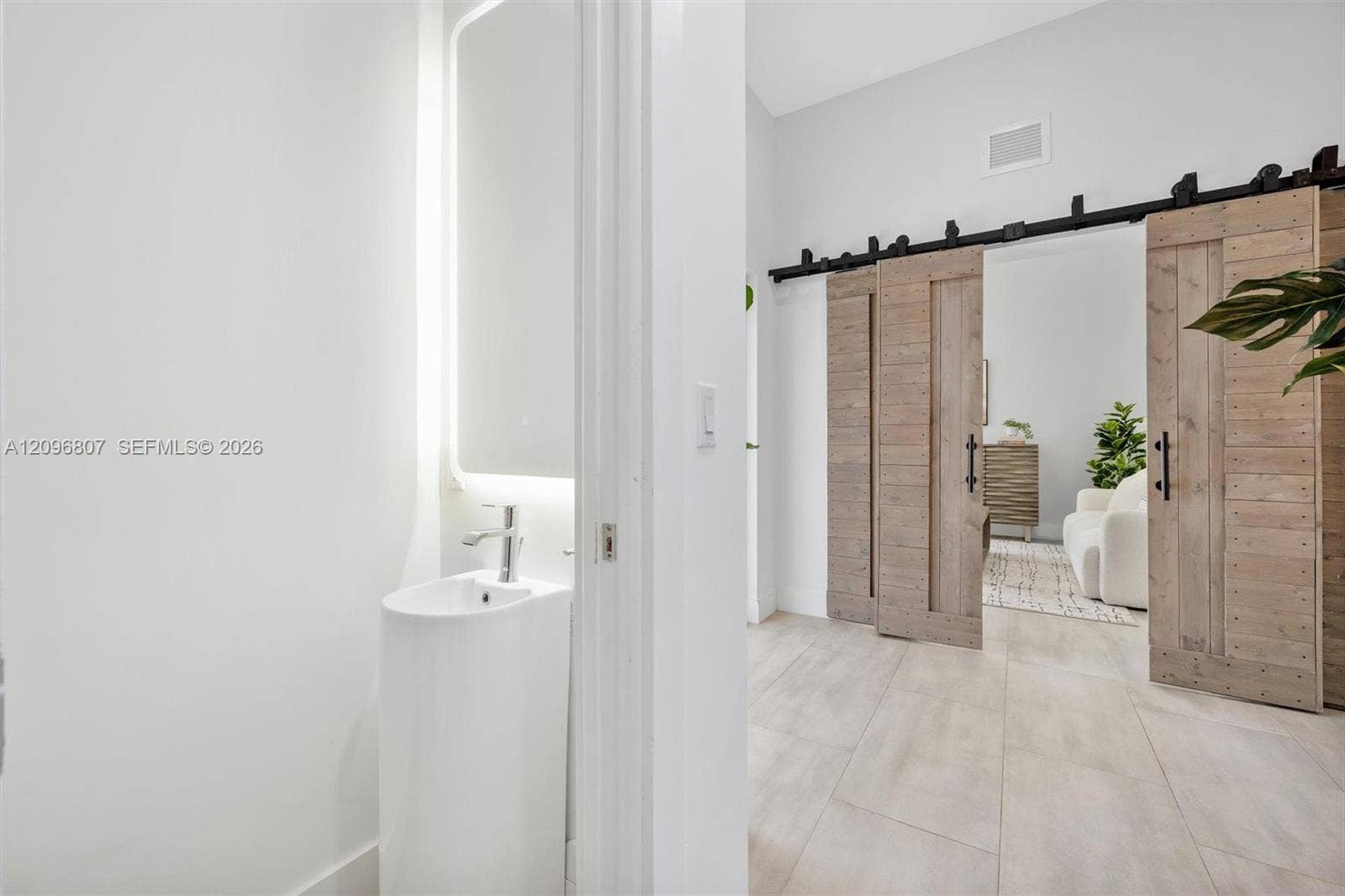 Pedestal sink with a single-handle chrome faucet, LED-backlit mirror, large-format tile flooring, and wood-finish sliding barn doors with matte black hardware