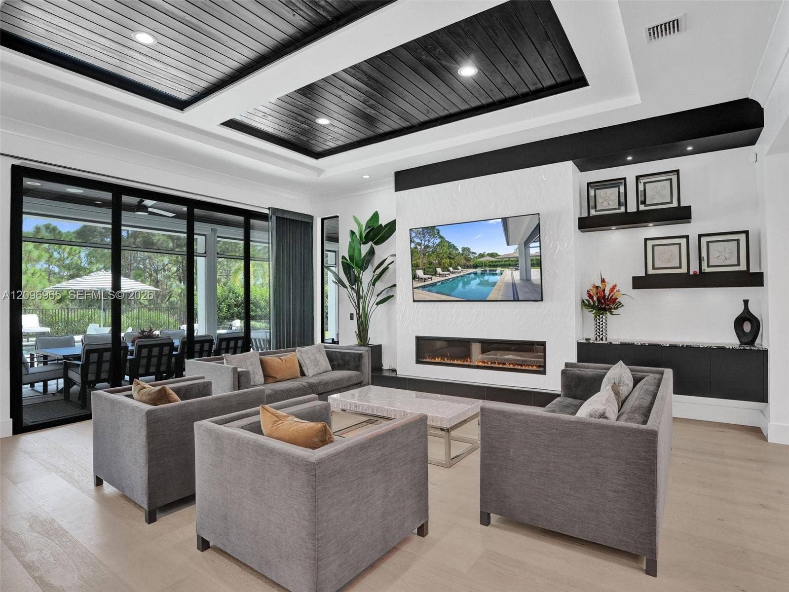10157 SE Sandpine Lane, Hobe Sound, FL 33455 Tray ceiling with dark wood-finish plank detailing, linear electric fireplace, built-in display shelving, wood-finish flooring, and multi-panel sliding glass doors