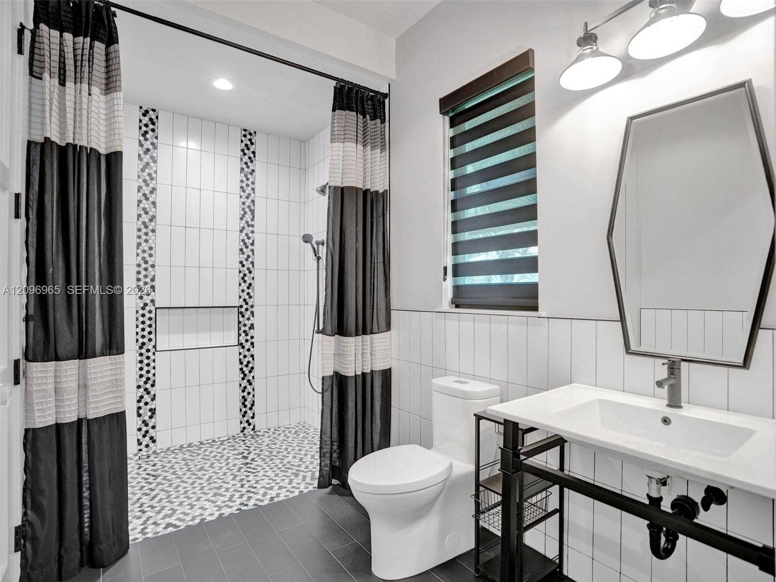 10157 SE Sandpine Lane, Hobe Sound, FL 33455 Bathroom featuring a shower stall with mosaic tile accents and a built-in recessed niche, complemented by a pedestal-style sink with a hexagonal mirror and matte black hardware