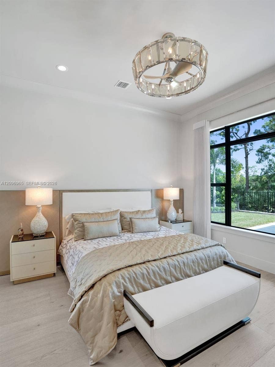 10157 SE Sandpine Lane, Hobe Sound, FL 33455 Light-filled room featuring wood-finish flooring, a custom upholstered wall panel, large black-framed windows, and a decorative circular crystal ceiling fan fixture