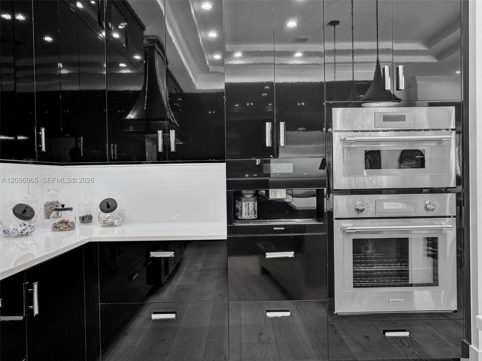 10157 SE Sandpine Lane, Hobe Sound, FL 33455 High-gloss black cabinetry featuring minimalist hardware, stainless steel wall-mounted oven and microwave, white solid-surface countertops, built-in coffee system, and wood-finish flooring