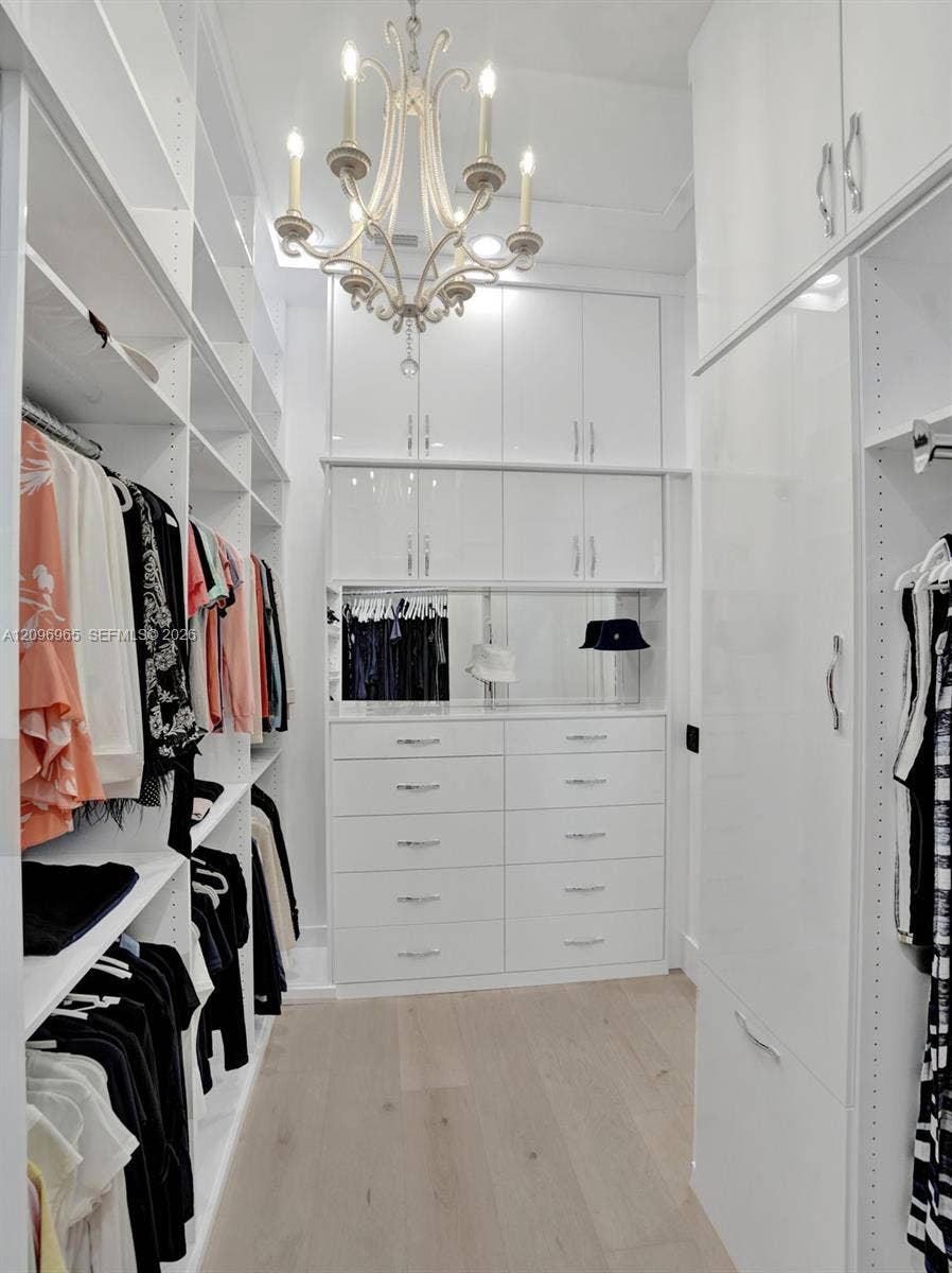 10157 SE Sandpine Lane, Hobe Sound, FL 33455 Custom walk-in closet featuring extensive white floor-to-ceiling cabinetry, integrated drawer banks, a central mirrored backsplash, and a multi-arm chandelier on wood-finish flooring