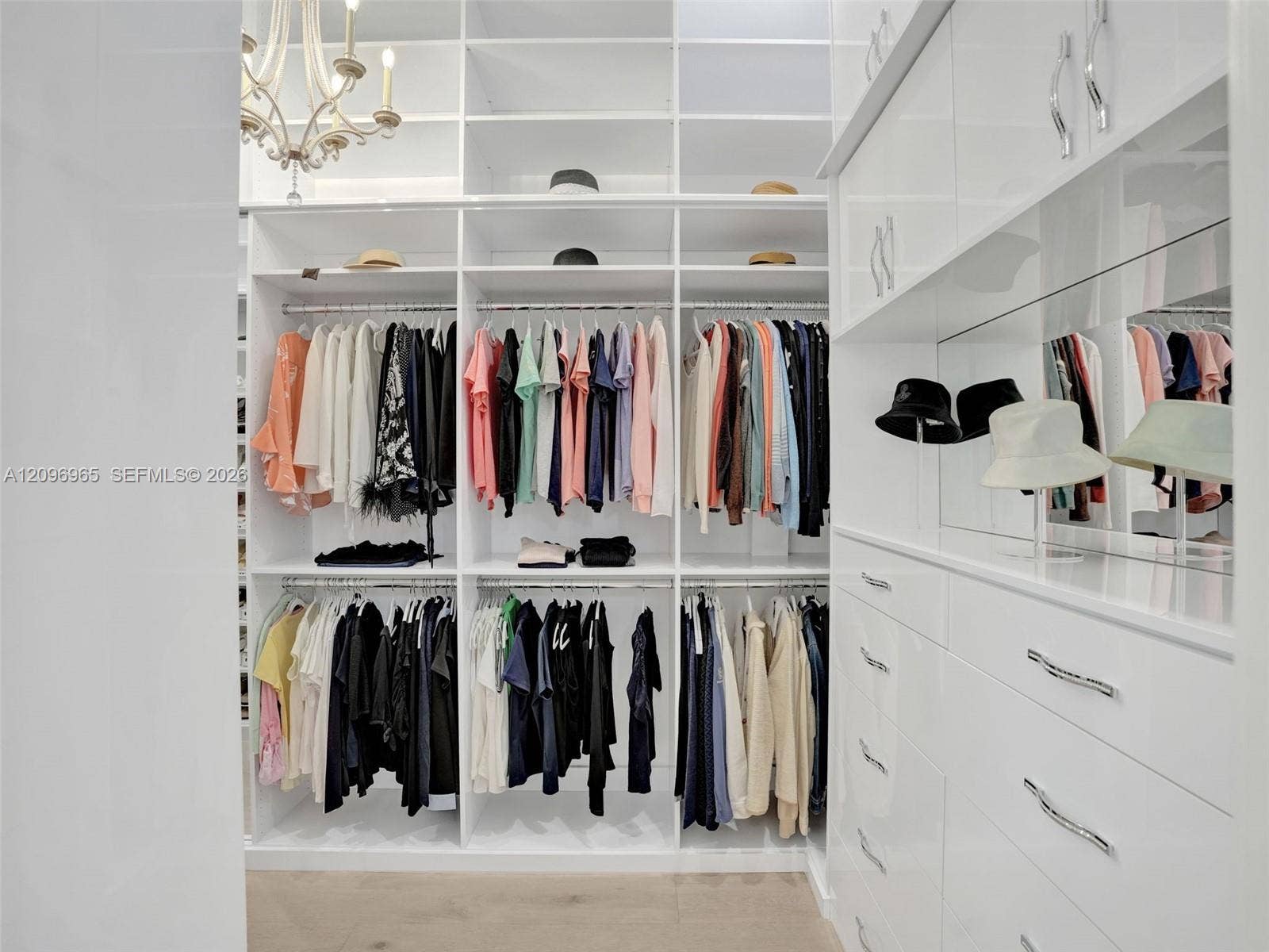 10157 SE Sandpine Lane, Hobe Sound, FL 33455 Custom walk-in closet featuring white floor-to-ceiling built-in cabinetry, polished chrome hardware, integrated mirror panels, and a decorative overhead chandelier
