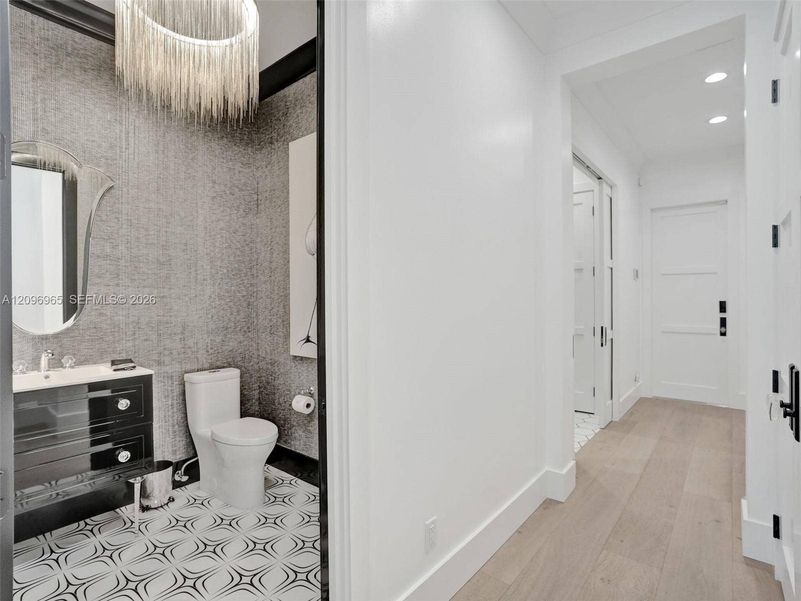 10157 SE Sandpine Lane, Hobe Sound, FL 33455 Powder room featuring a high-contrast vanity with integrated basin, textured wall covering, custom fringed chandelier, patterned tile flooring, and wood-finish flooring in the hallway