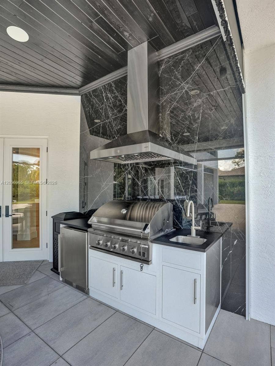 10157 SE Sandpine Lane, Hobe Sound, FL 33455 Outdoor kitchen featuring a stainless steel grill, under-counter refrigeration, prep sink, dark stone backsplash, and white cabinetry