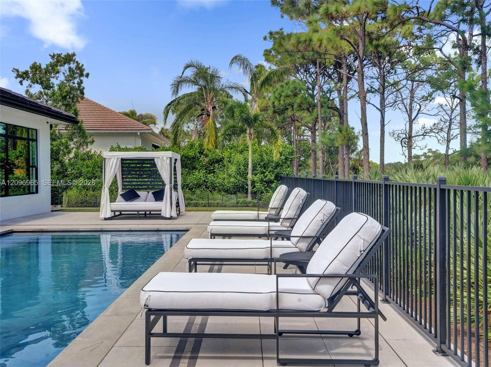 10157 SE Sandpine Lane, Hobe Sound, FL 33455 In-ground swimming pool with stone-tiled decking and black metal perimeter fencing, featuring mature pine tree landscaping