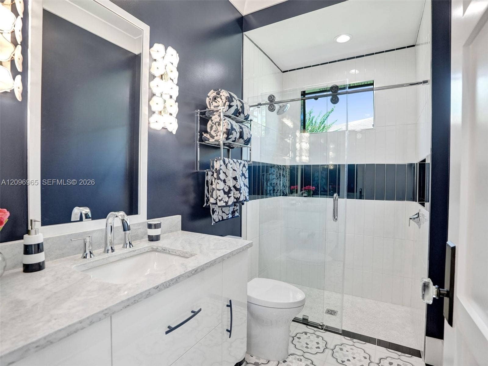 10157 SE Sandpine Lane, Hobe Sound, FL 33455 Bathroom featuring a vanity with a light-toned stone countertop, white cabinetry with matte black hardware, a glass-enclosed shower with tiled walls, and patterned floor tile