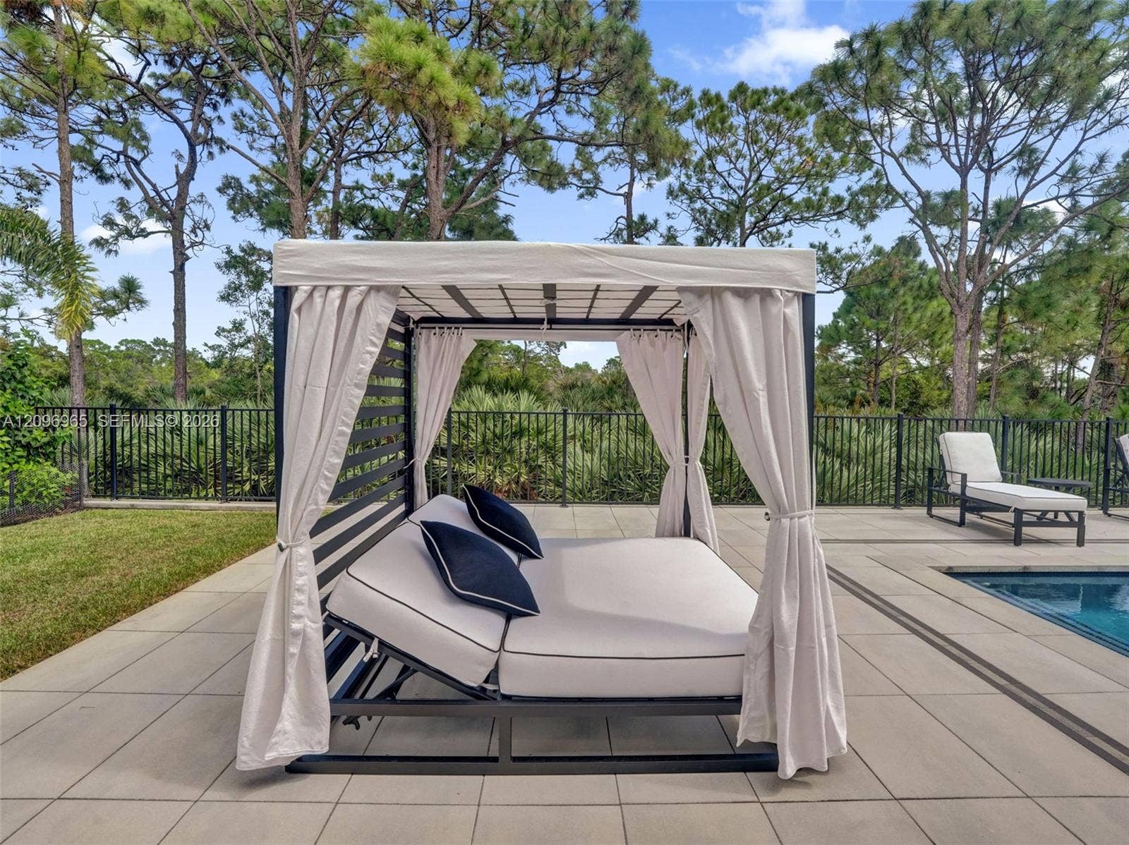 10157 SE Sandpine Lane, Hobe Sound, FL 33455 Spacious paver patio featuring an in-ground pool, black metal perimeter fencing, and mature pine tree surroundings