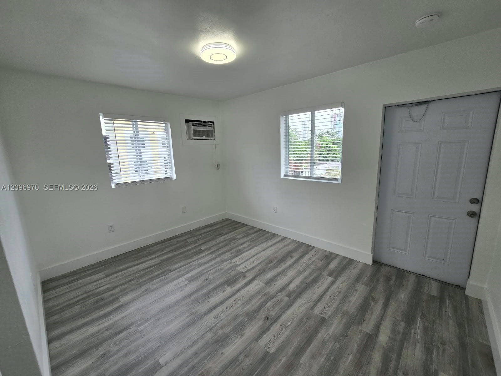 535 NW 7th Street, #12, Miami, Miami, FL 33136 Interior space featuring wood-finish flooring, white walls, dual windows with blinds, a flush-mount ceiling light fixture, and a six-panel entry door