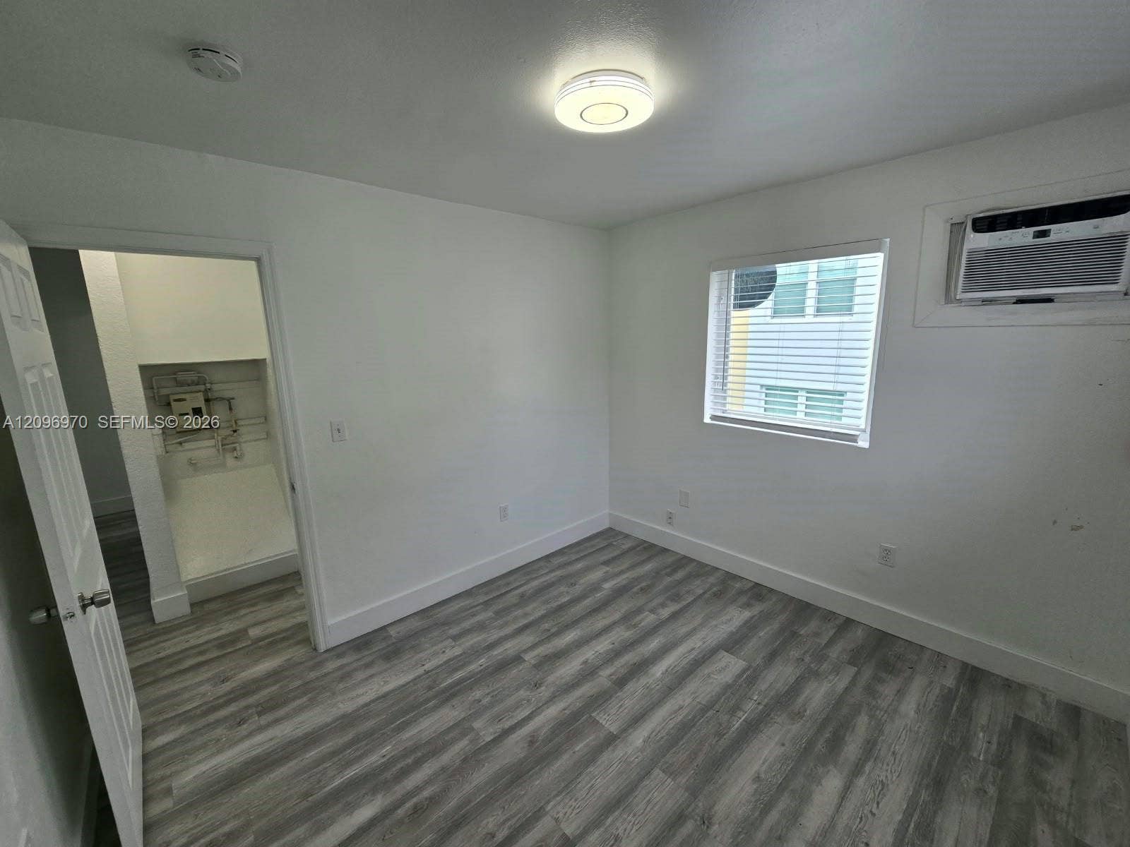 535 NW 7th Street, #12, Miami, Miami, FL 33136 Neutral-toned room featuring wood-finish flooring, a single-hung window with installed blinds, a wall-mounted air conditioning unit, and a flush-mount ceiling light