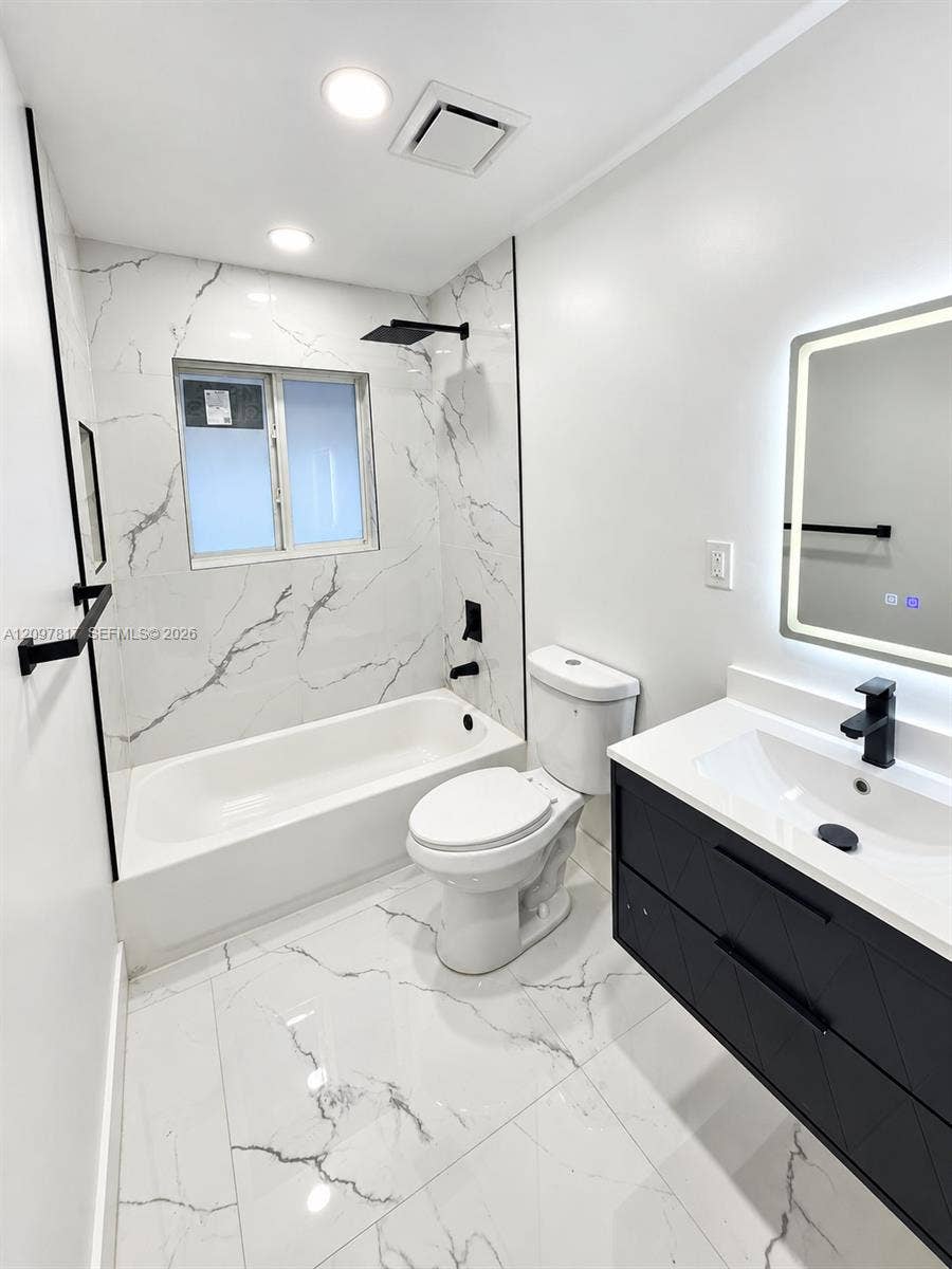 Bathroom featuring large-format stone-patterned tile flooring and wall surrounds, dark finish floating vanity with white countertop, matte black fixtures, and a wall-mounted LED mirror