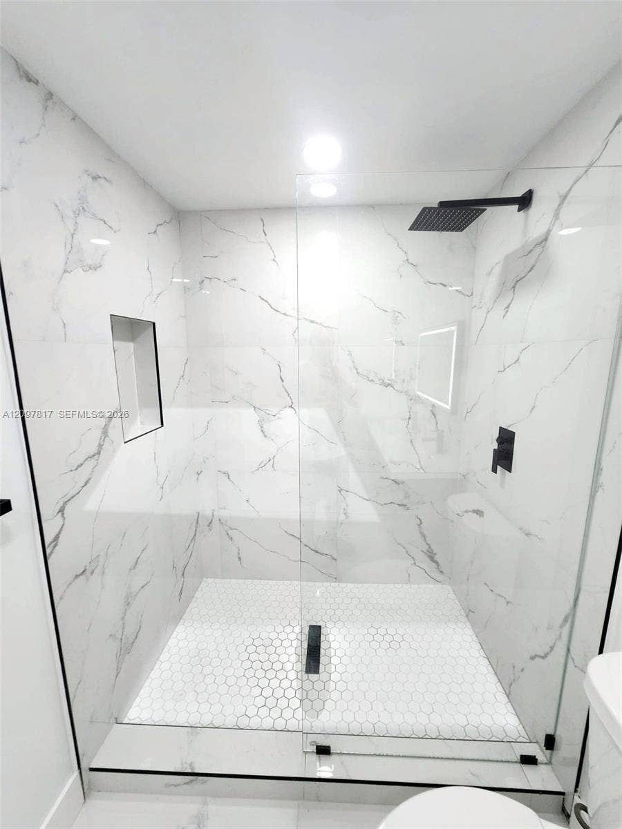 Walk-in shower featuring large-format white stone-finish wall tile, hexagonal floor tiling, a frameless glass enclosure, a built-in recessed niche, and matte black plumbing fixtures