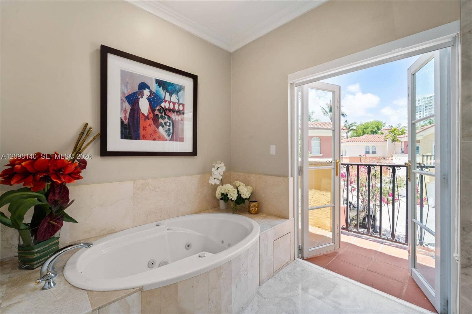 Jetted soaking tub with stone-tiled surround, crown molding, and double-door access to a private balcony with wrought-iron railing