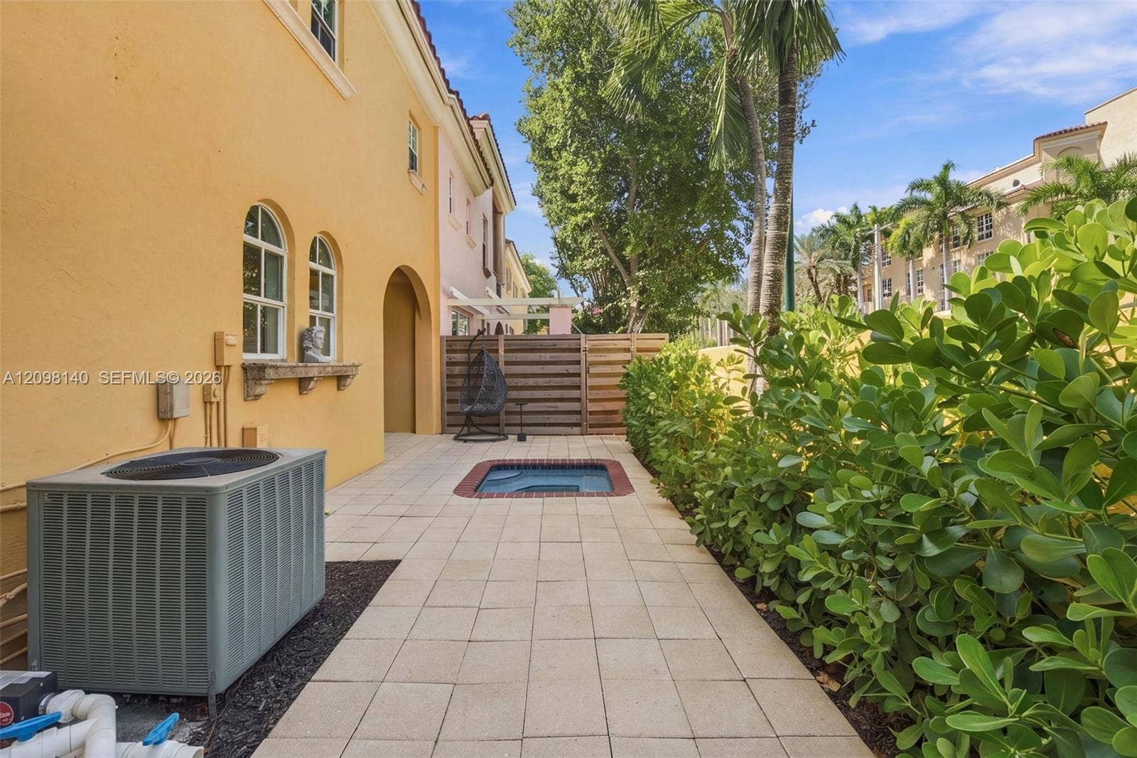 Exterior patio featuring light-toned stone tile flooring, an in-ground spa with brick coping, horizontal wood privacy fencing, and lush perimeter shrubbery