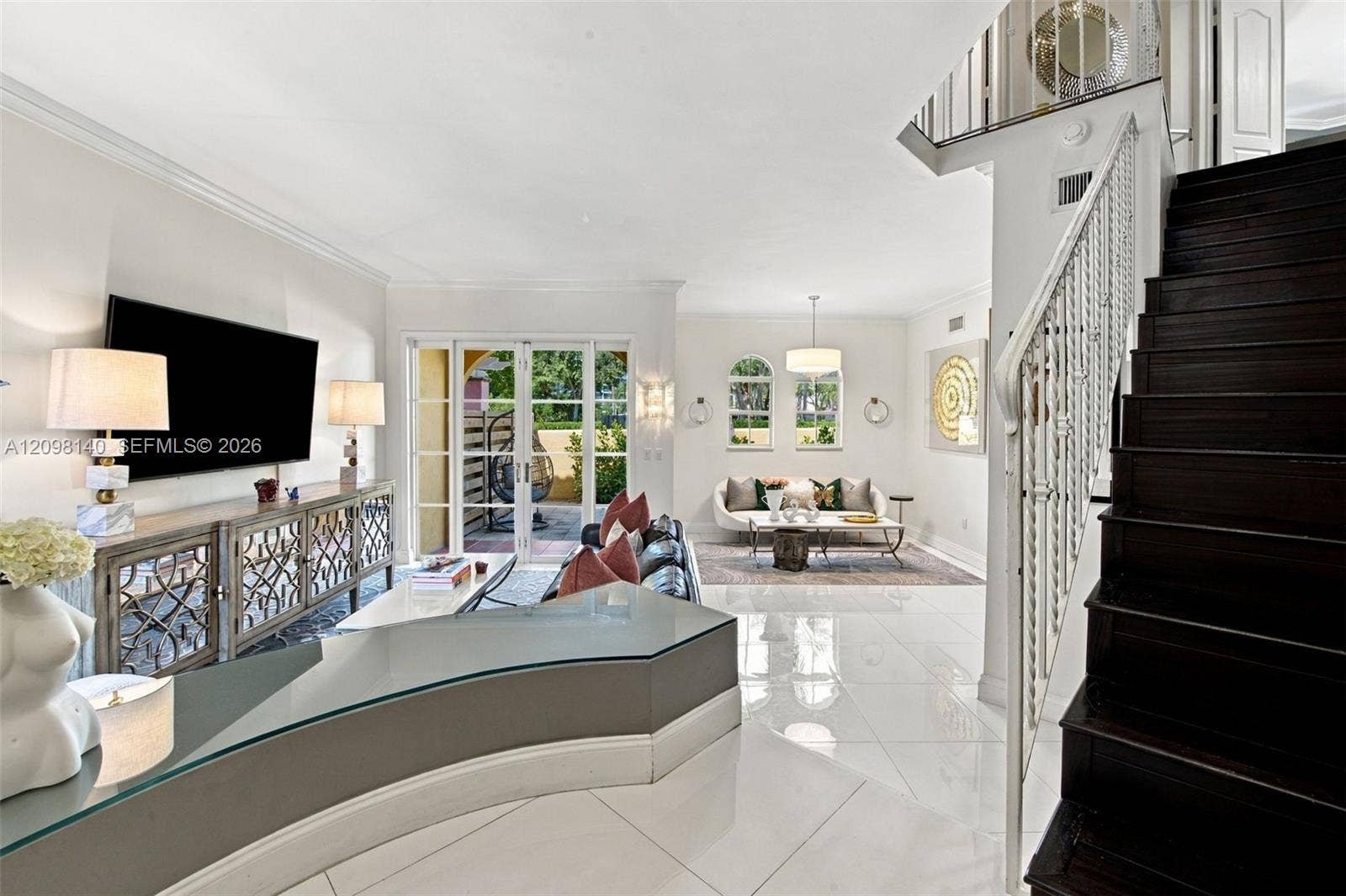 Open-concept living space featuring high-gloss tile flooring, an architectural curved built-in desk with glass surface, a dark-stained wood staircase with decorative metal railing, and multi-pane glass French doors