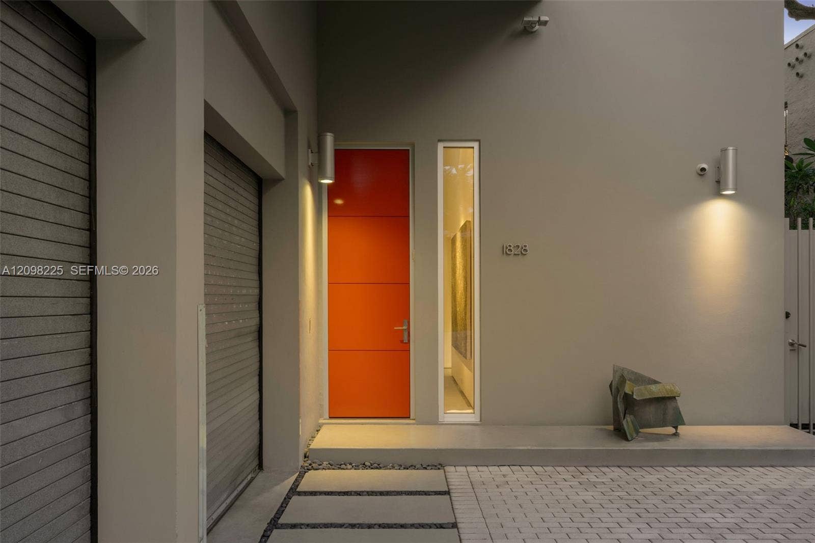 Modern architectural entry featuring a vibrant orange slab door, vertical transom window, dual cylinder wall-mounted exterior light fixtures, and a paved walkway with concrete pavers