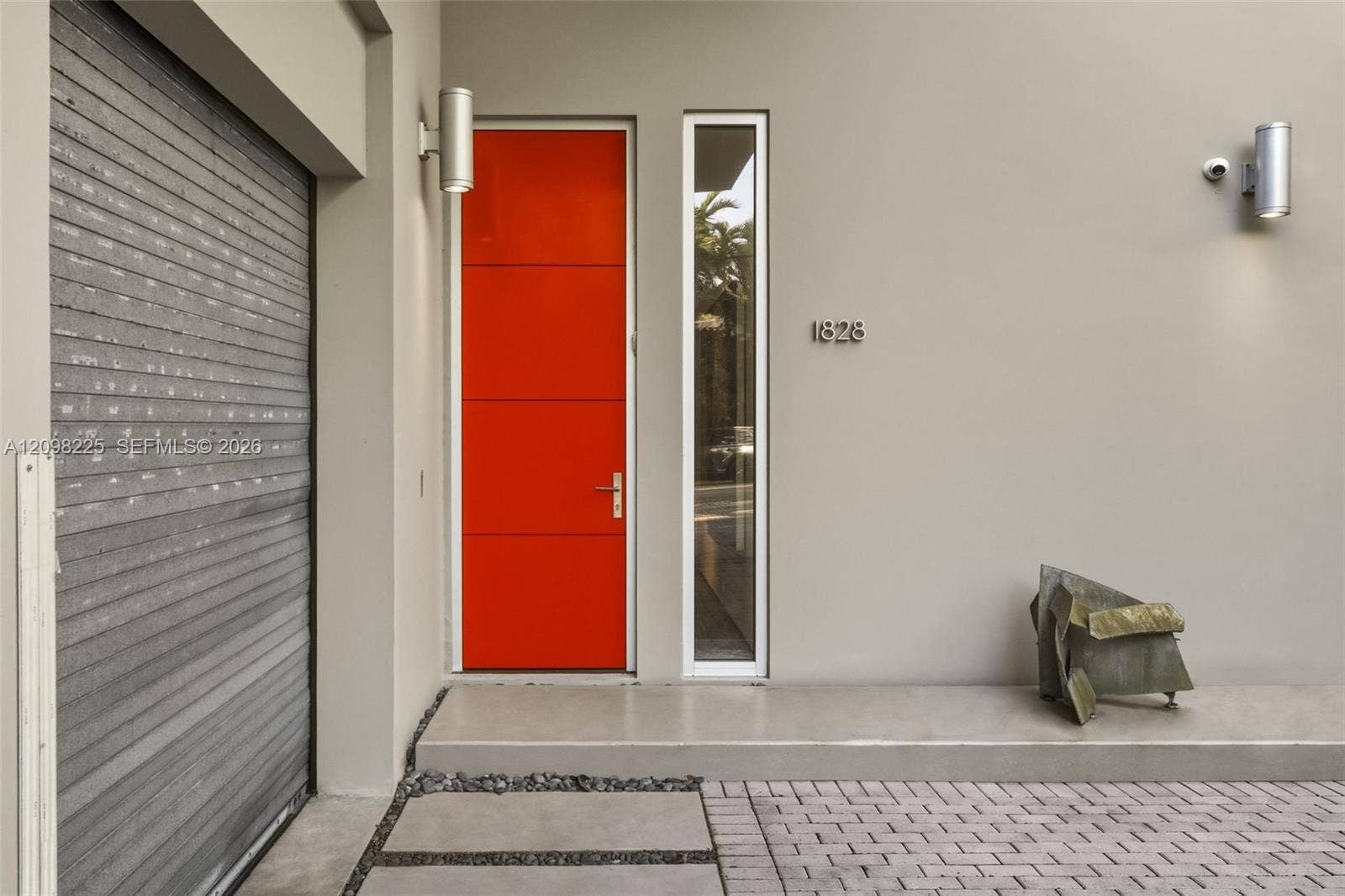 Modern exterior entry featuring a high-contrast orange door, tall side-light window, metallic wall-mounted light fixtures, and a paved brick driveway