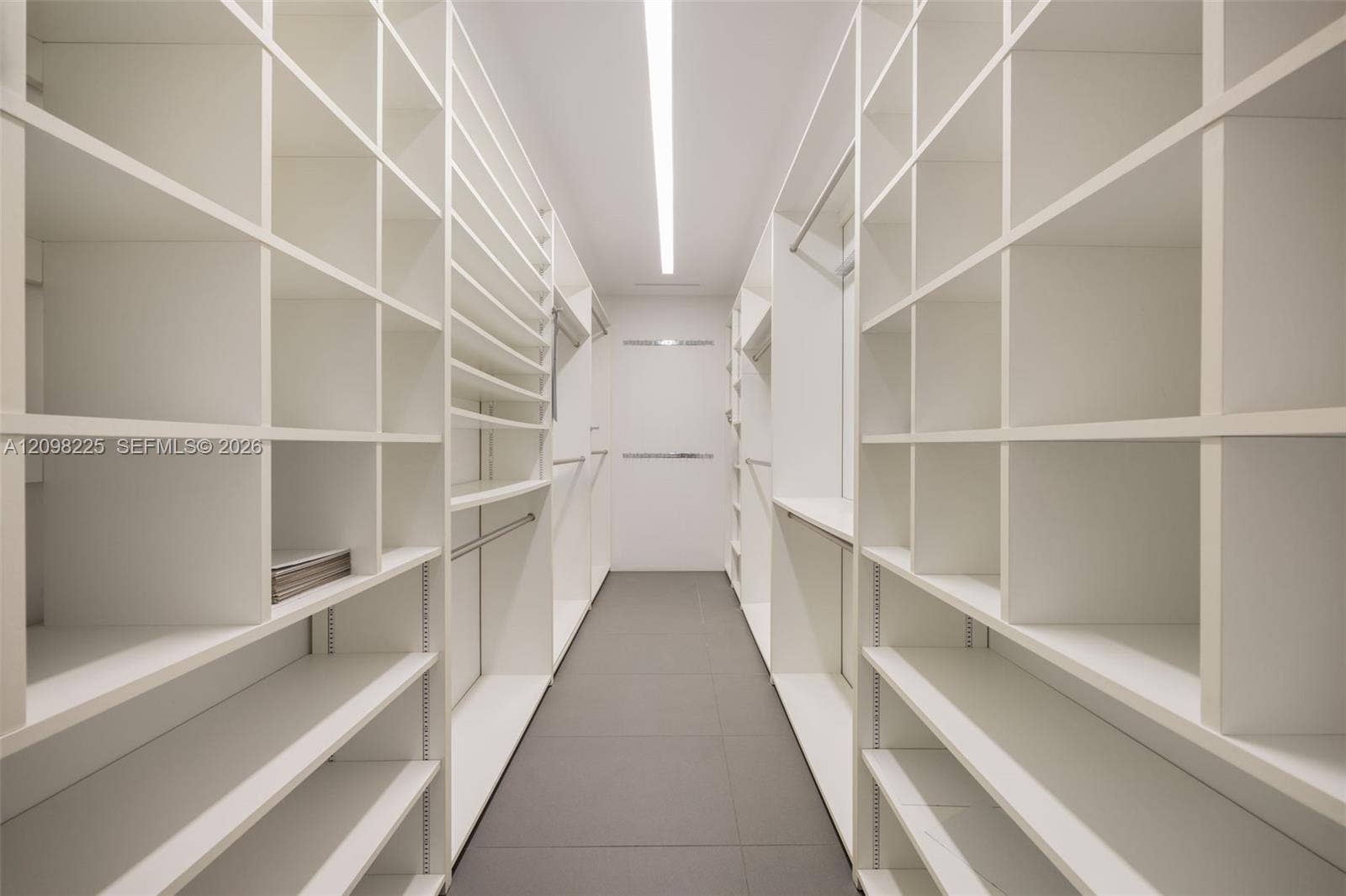 Custom walk-in storage featuring floor-to-ceiling white shelving units, integrated hanging rods, and large-format tile flooring with linear overhead lighting