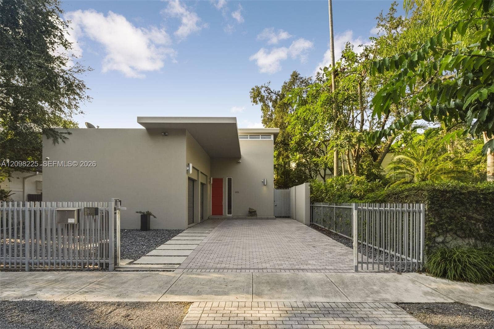 Modern architectural exterior featuring smooth stucco siding, a flat roofline with an extended entryway overhang, gated paver driveway, and vertical metal privacy fencing