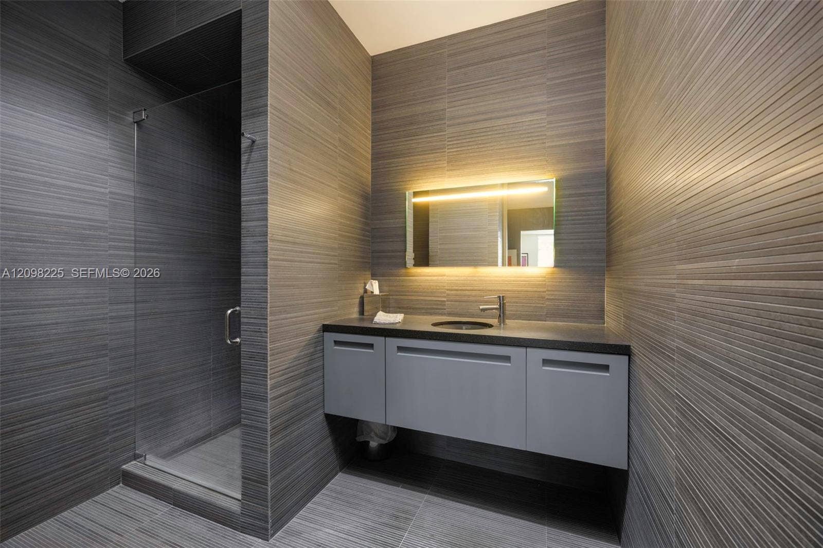 Modern bathroom featuring floor-to-ceiling linear-textured wall tile, a floating vanity with a solid surface countertop, a built-in backlit mirror, and a glass-enclosed shower stall