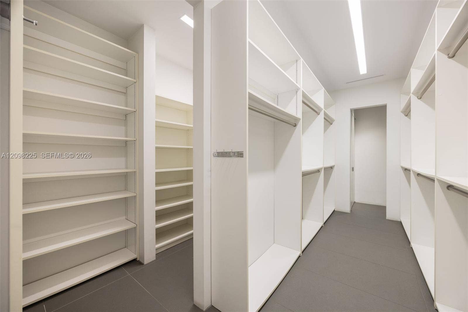 Custom walk-in closet featuring floor-to-ceiling white shelving, integrated hanging rods, minimalist cabinetry, and large-format tile flooring with recessed linear lighting