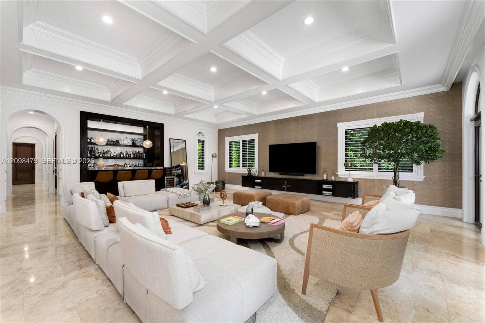 Coffered ceiling with recessed lighting, integrated wet bar, large windows with white shutters, wall-mounted media console, and stone tile flooring