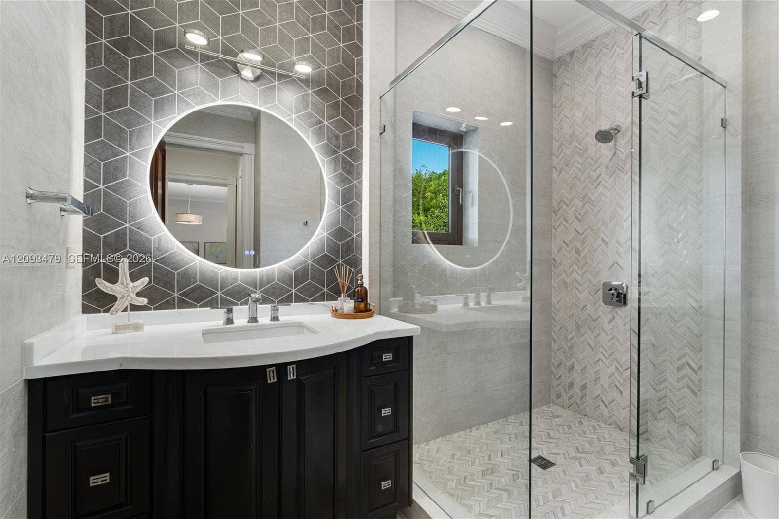Vanity with integrated sink and chrome fixtures, illuminated round mirror, geometric wall tile, glass-enclosed shower, and herringbone tile shower surround